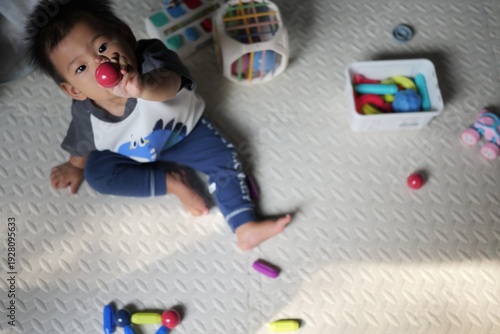 Asian toddler boy sitting on a soft play mat indoors, holding a red toy ball and looking up at the camera. 