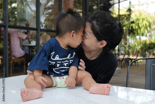 Asian mother gently touching foreheads with her baby boy while sitting at an outdoor café.