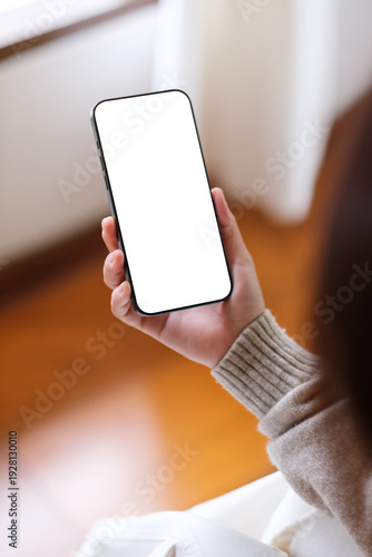 Vertical mockup image of a woman holding and using mobile phone with blank desktop screen at home