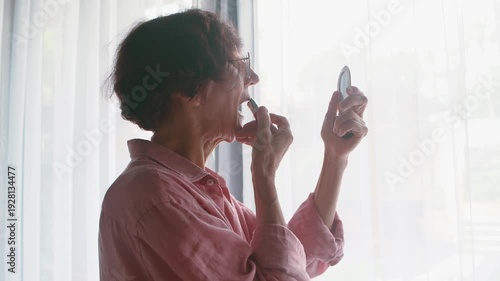 Close-up of elderly caucasian woman with eyeglasses doing makeup holding lipstick and small mirror in her hand