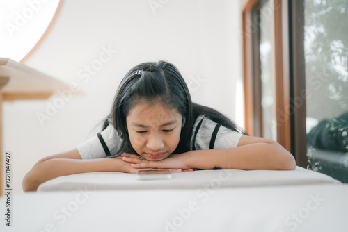 A young girl is resting her chin on her arms while looking thoughtfully at a smartphone placed on a table in a bright and quiet room.