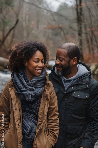 Two people stand close together by a river in the woods during autumn. They smile at each other while enjoying the moment. Trees surround them, and the atmosphere feels warm from their connection