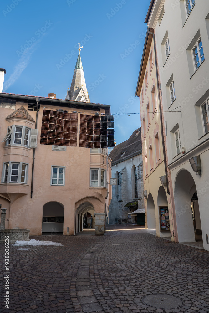 Fototapeta premium A cobblestone lane in Brixen, Italy, winds by pastel arcades and closed shops. A canopy of dark bottles spans the street. The cathedral spire rises in soft winter light.