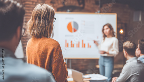 A group of professionals attentively listens to a woman presenting data charts on a whiteboard during a business meeting.