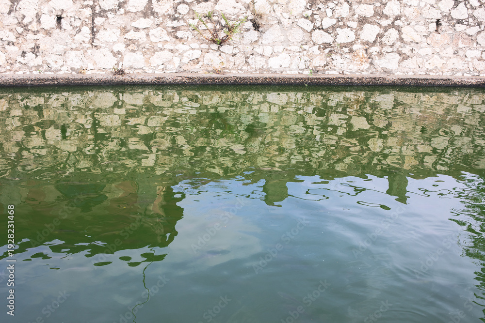 Naklejka premium Stone wall reflects on surface of calm greenish water, creating a textured and abstract mirrored pattern. Ripples distort the reflection, blending the wall rough tones