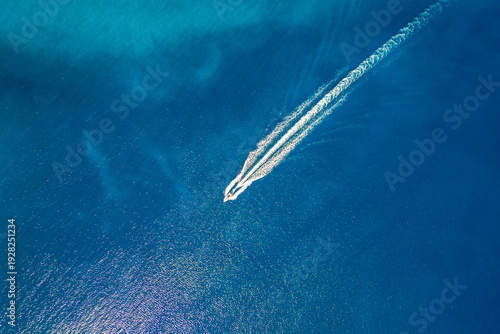 High-angle aerial shot of a single powerboat creating a sharp white wake through deep blue water, capturing the essence of speed, travel, and tranquility on a vast, sunlit open ocean surface.