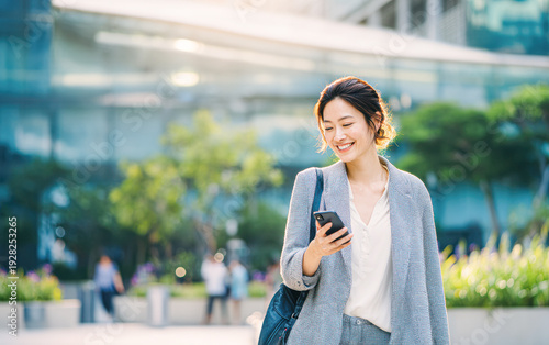 A young woman smiles while looking at her phone outdoors in an urban setting, dressed in business casual attire with a bag over her shoulder. Free Download A young woman smiles while looking at her phone outdoors in an urban setting, dressed in business casual attire with a bag over her shoulder. - GFXLoad