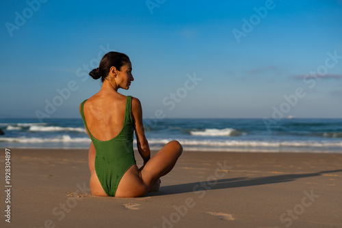 Εκτύπωση καμβά Fit woman in a green textured swimsuit sitting on the beach in a relaxed pose, looking at the ocean, enjoying peaceful morning solitude and the sound of the waves