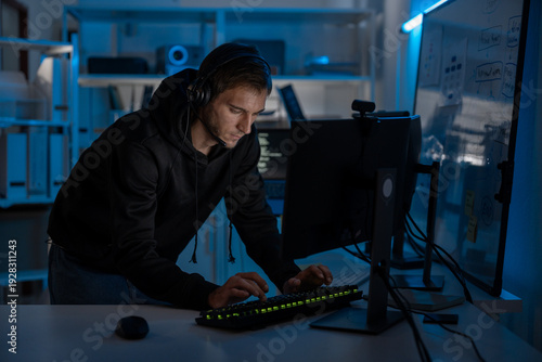 Male programmer and developer focused on computer screen at night, coding digital systems and ensuring cyber security. Freelancer with headset working overtime under blue light in office.