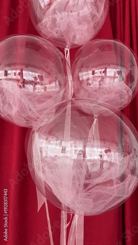 Four transparent bobo balloons filled with soft pink tulle fabric, hanging against a rich red velvet curtain. Elegant and trendy festive decoration