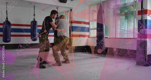 Grappling two mid-adult men sparring at combat gym with hanging bags, in camo and tactical boots