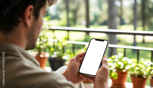 Hand holding mobile smartphone with blank white screen mockup, man relaxing on sunny balcony with potted plants, over shoulder view, digital app display, nature summer garden
