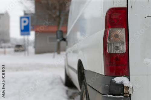 White van parked on snowy curb showing taillight, bumper and parking sign, distant buildings blurred in cold morning, salt stains and slush around wheel, last mile delivery vehicle waiting