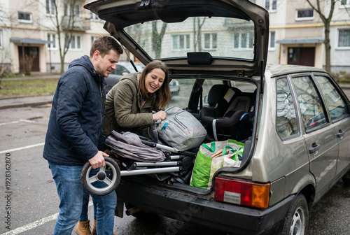 Young parents loading stroller and baby gear into car trunk, preparing for family outing in residential parking lot.