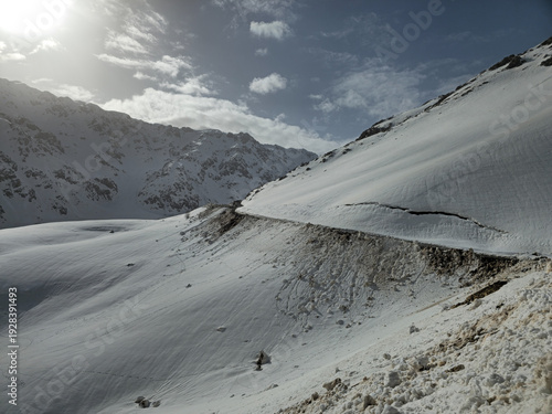Fényképezés Sunlit snowy mountain road winding across steep winter slopes, highlighting the dramatic contrast of bright sunlight, deep shadows, and untouched high‑altitude terrain