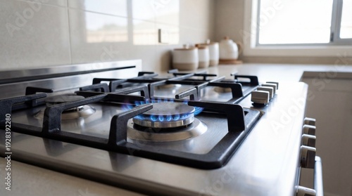 Stainless steel gas stove with blue flame ignited, modern kitchen setting featuring white cabinets and ceramic containers on countertop near window