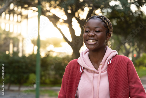 African American teen in pink hoodie walking, smiling on paved path near green lamppost, copy space