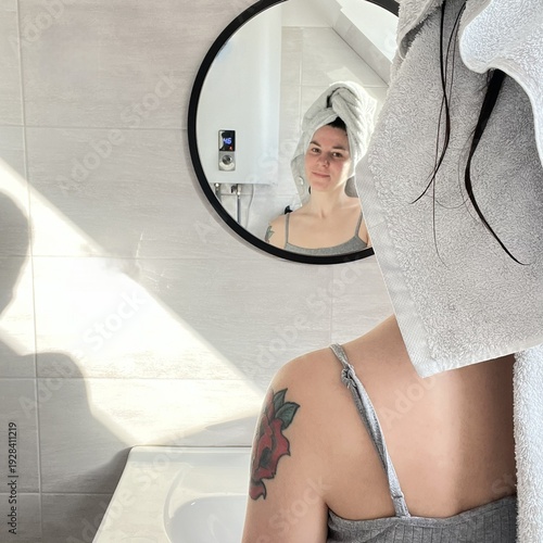 Tableau sur toile Young woman with towel on head looking at herself in bathroom mirror
