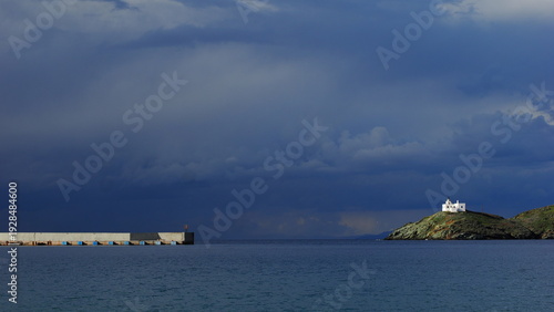 Photos White lighthouse standing on a rocky promontory beneath dark dramatic storm clouds in the Aegean Sea, Greece