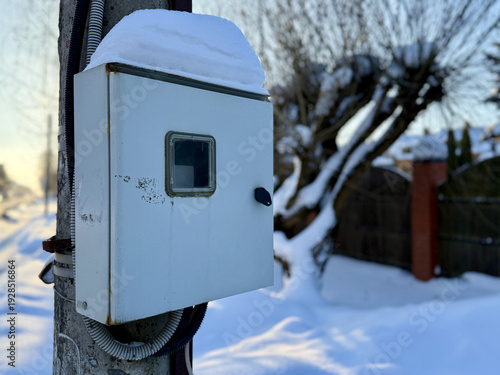 Snow covered electrical panel on pole beside cottage under soft dawn light, conduit and cables wrapped along concrete pole, rust