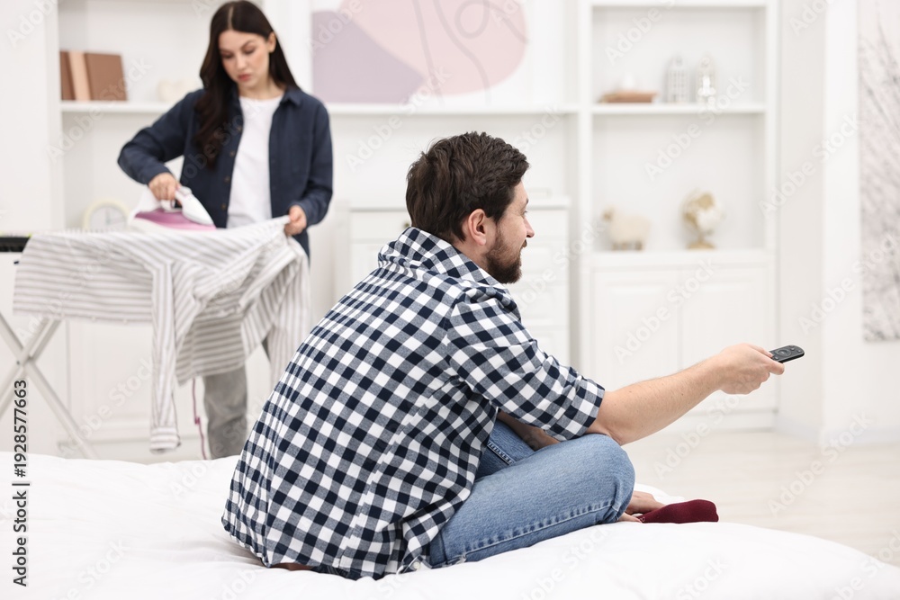 Obraz premium Gender inequality concept. Man with remote control sitting on bed while his wife ironing clothes indoors, selective focus