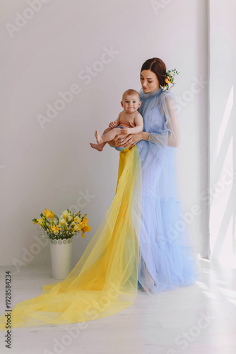 Mother holding baby with flowing yellow and blue fabric, woman in pastel tulle dress in bright minimal interior, motherhood lifestyle concept