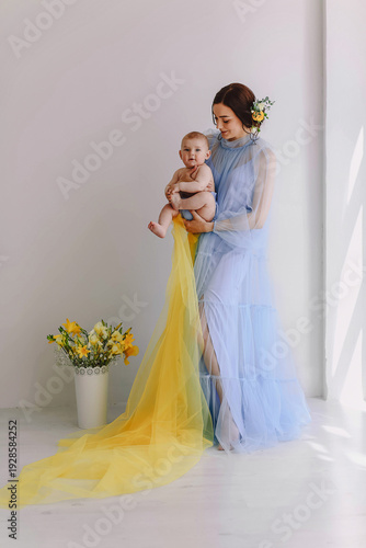 Happy mother holding infant child in light blue tulle dress, emotional bonding moment in bright home interior