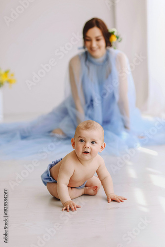 Curious crawling baby in blue bloomers with smiling mother in light blue tulle dress in soft bright interior