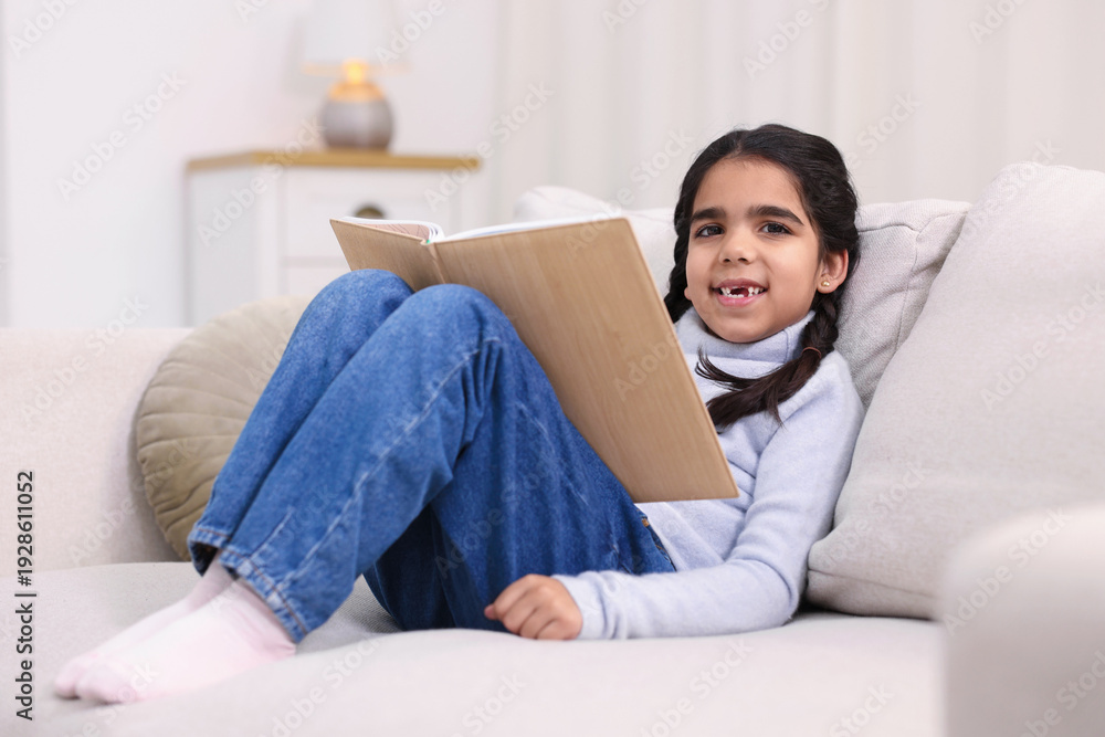 Naklejka premium Cute little girl reading book on couch at home