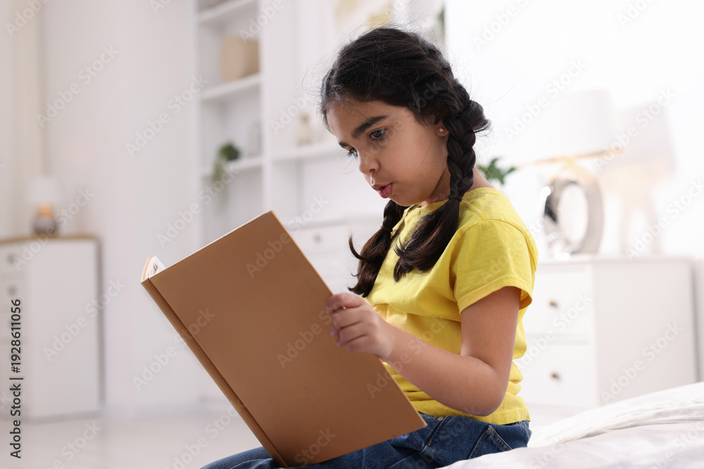 Naklejka premium Cute little girl reading book on bed at home