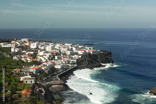 Coastal village of Las Aguas in San Juan de la Rambla, Tenerife. White houses sit atop dark volcanic cliffs as Atlantic waves crash against the rugged shoreline under a hazy sky