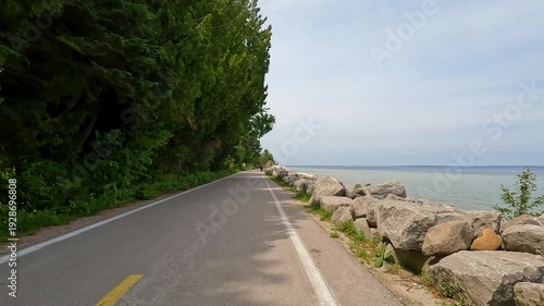 Slow-motion POV biking M-185 on Mackinac Island, MI. The paved coastal road stretches ahead with cyclists and pedestrians, flanked by a rocky Lake Huron shore and green forest.