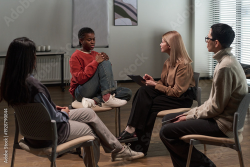 Group of adults and one Black gen Z girl sitting in circle engaging in discussion during support group meeting, female facilitator holding clipboard listening attentively
