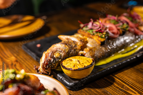 Photography Whole grilled fish presented on a black serving platter with a charred lemon, fr