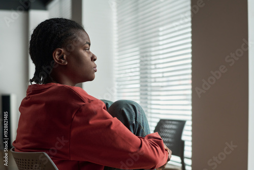 Black woman sitting by window gazing outside with knees drawn up, wearing casual clothing, natural light streaming through blinds, depressed expression visible on face Free Download Black woman sitting by window gazing outside with knees drawn up, wearing casual clothing, natural light streaming through blinds, depressed expression visible on face - GFXLoad