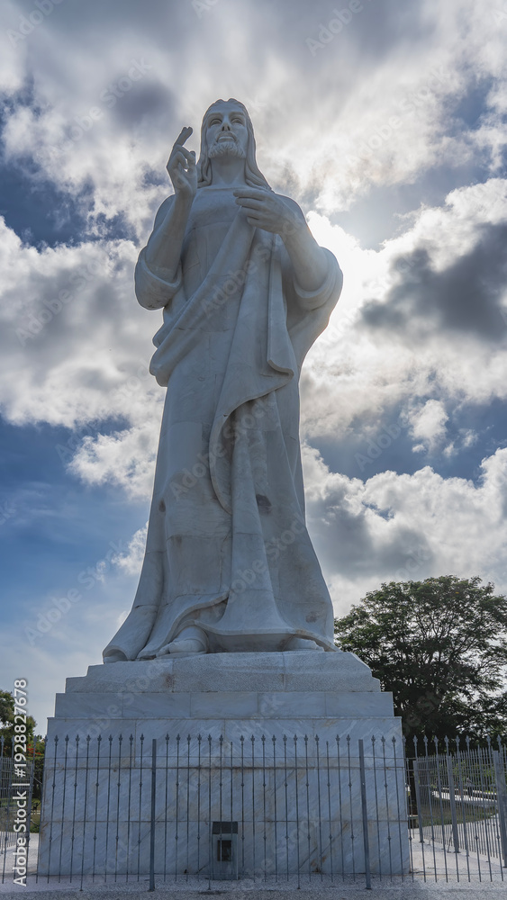 Fototapeta premium The famous sculpture of Christ of Havana. A tall statue made of white carrara marble against a background of blue sky and clouds. The pedestal is enclosed by a latticed fence. Cuba. Havana.