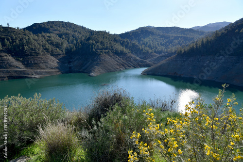 A high-angle landscape of the turquoise Lefkara dam reservoir surrounded by pine-covered mountains and rocky terrain under a bright Mediterranean sun.