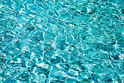 Bright sunlight illuminates the clear blue water surface, forming dynamic ripple patterns. This vibrant scene shows calm, refreshing liquid movement in a swimming pool under the warm sun.