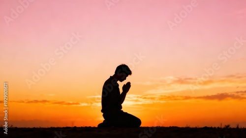 Man kneels in prayer with hands pressed together against a beautiful orange and pink sunset sky at dusk, creating a peaceful silhouette of faith and devotion.
