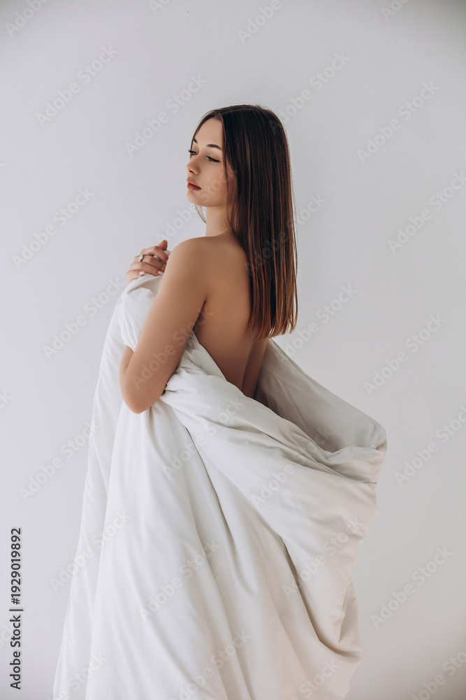 Fototapeta premium Beauty portrait of a model, a beautiful brunette girl with straight long hair dressed in a long white blanket on a white background