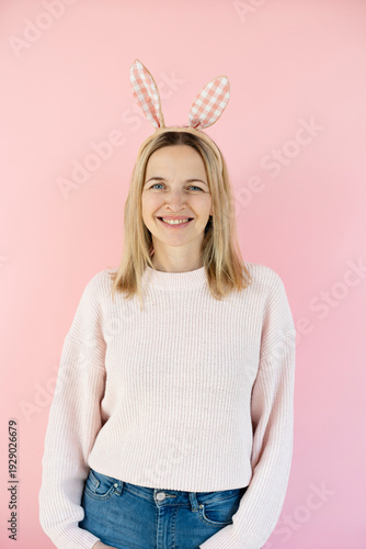 Pretty middle-aged blonde woman with bunny ears decoration standing in front of pink background