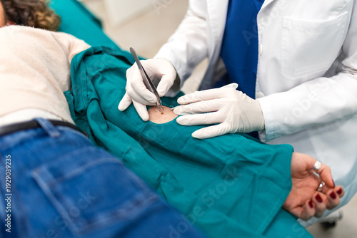A detailed macro view of a doctor performing a skin biopsy or mole removal on a patient's arm using surgical instruments. The physician wears white gloves and works over a sterile green medical drape.