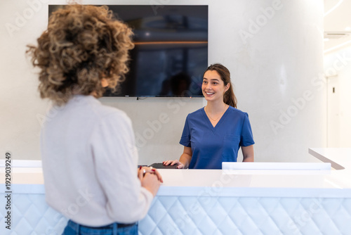 Fotografie Smiling receptionist in blue scrubs greeting patient at clinic front desk