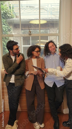 Four Diverse business people stand close together near a window, discussing content on a tablet while sharing ideas, feedback, and teamwork in a calm modern office environment.