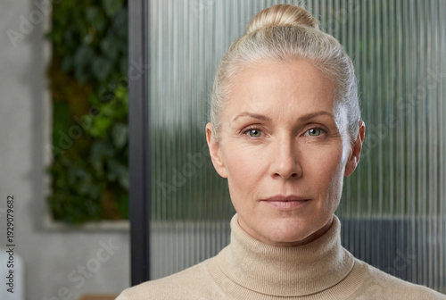 Portrait of a confident mature woman with grey hair in a bun wearing a beige turtleneck, standing in a modern office or clinic with a blurred green wall background.