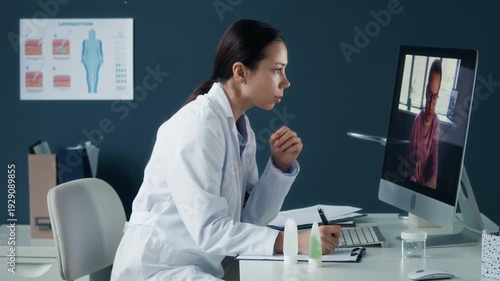 Female dermatologist conducting telemedicine consultation with patient on desktop computer in modern clinical office, eyelevel side view medium shot with medical workspace interior