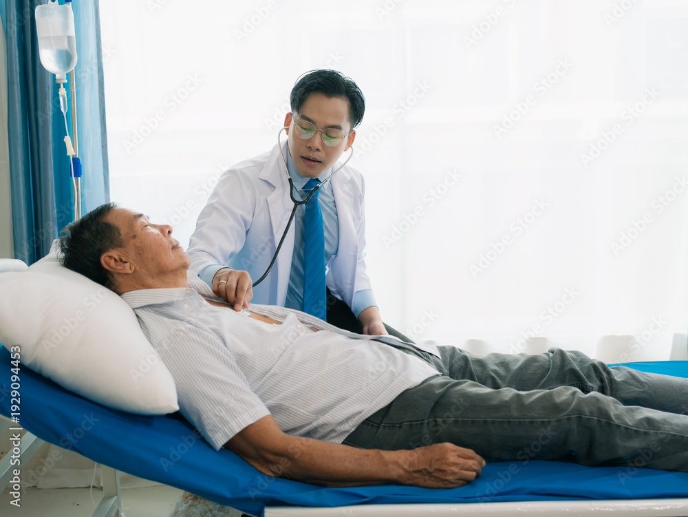 Fototapeta premium An Asian male doctor is examining an elderly patient with a stethoscope, conducting a routine health check. The male patient lies down for observation in the clinic.