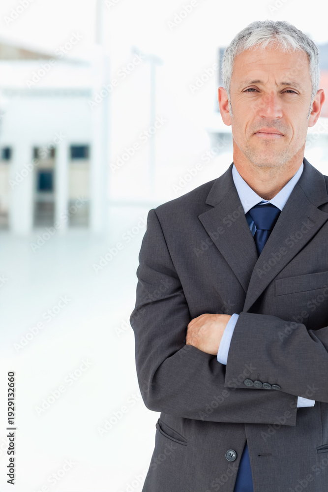Fototapeta premium Man standing with crossed arms facing forward in modern lobby wearing dark suit, copy space
