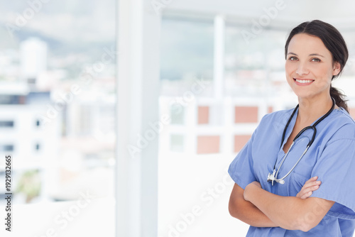 Fotografi Female clinician standing with arms crossed by clinic window in scrubs stethosco