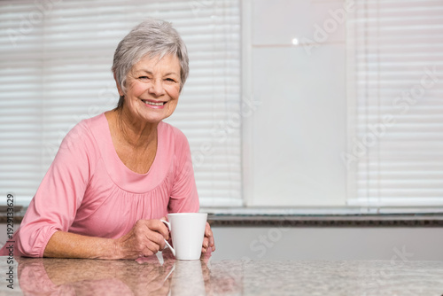 Senior woman leaning on granite counter holding mug in pink by kitchen window blinds, copy space